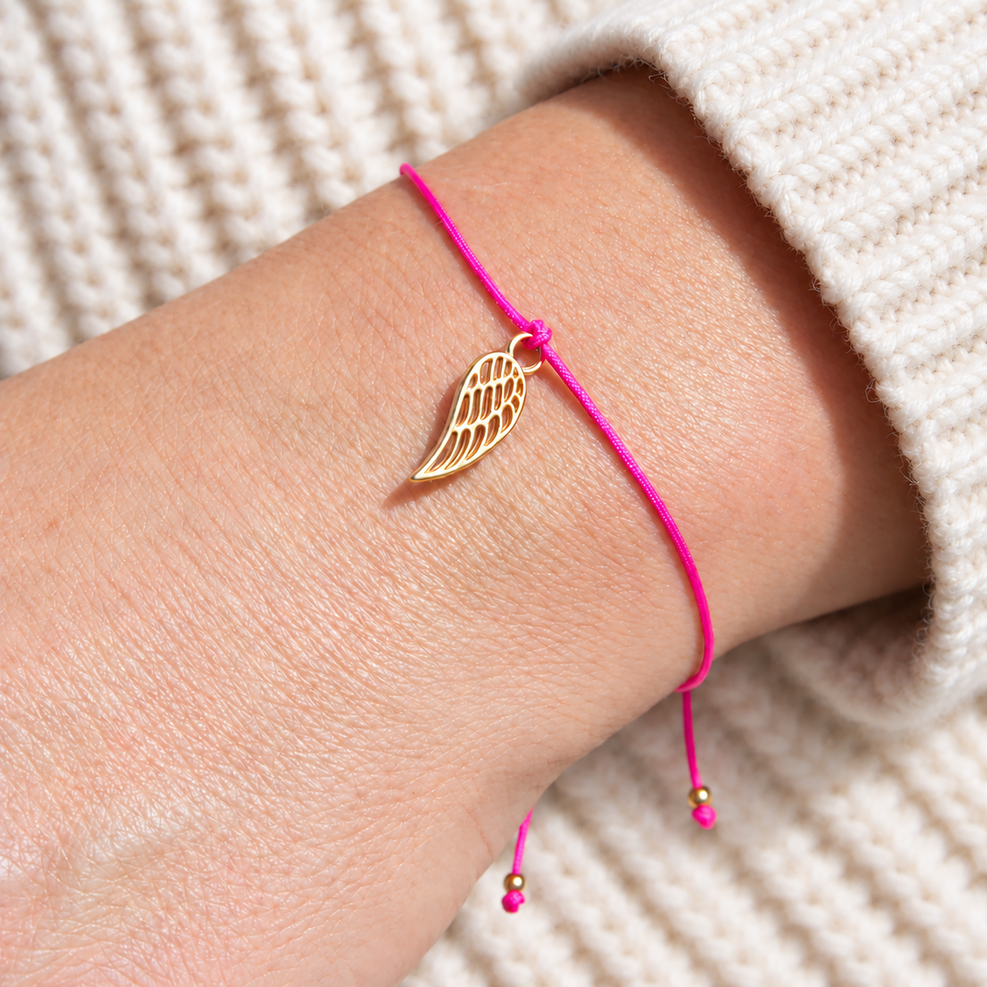 Schutzengel-Armband mit pinkem Band, goldenem Engelsflügel und kleinen goldenen Perlen an den Bandenden, am Handgelenk getragen.