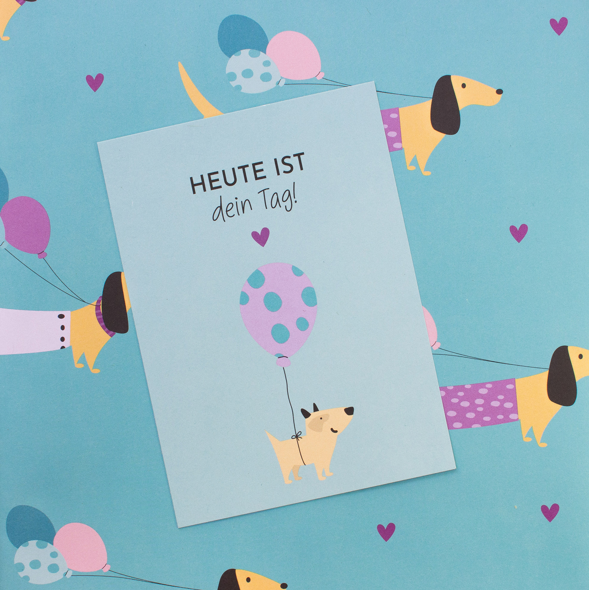Türkises Geschenkpapier mit Dackeln, Luftballons und Herzen, darauf eine Geburtstagskarte mit Hund und dem Spruch „Heute ist dein Tag!“.