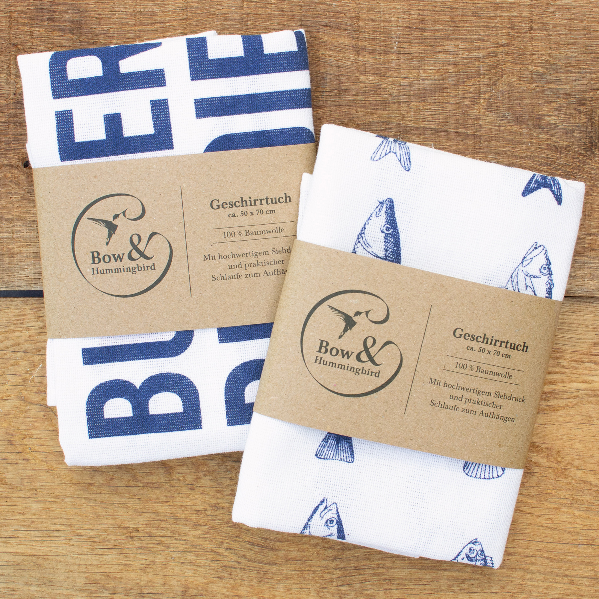 Gefaltetes Geschirrtuch „Butter bei die Fische“ mit blauem Schriftzug und Papierbanderole von Bow & Hummingbird auf Holz.