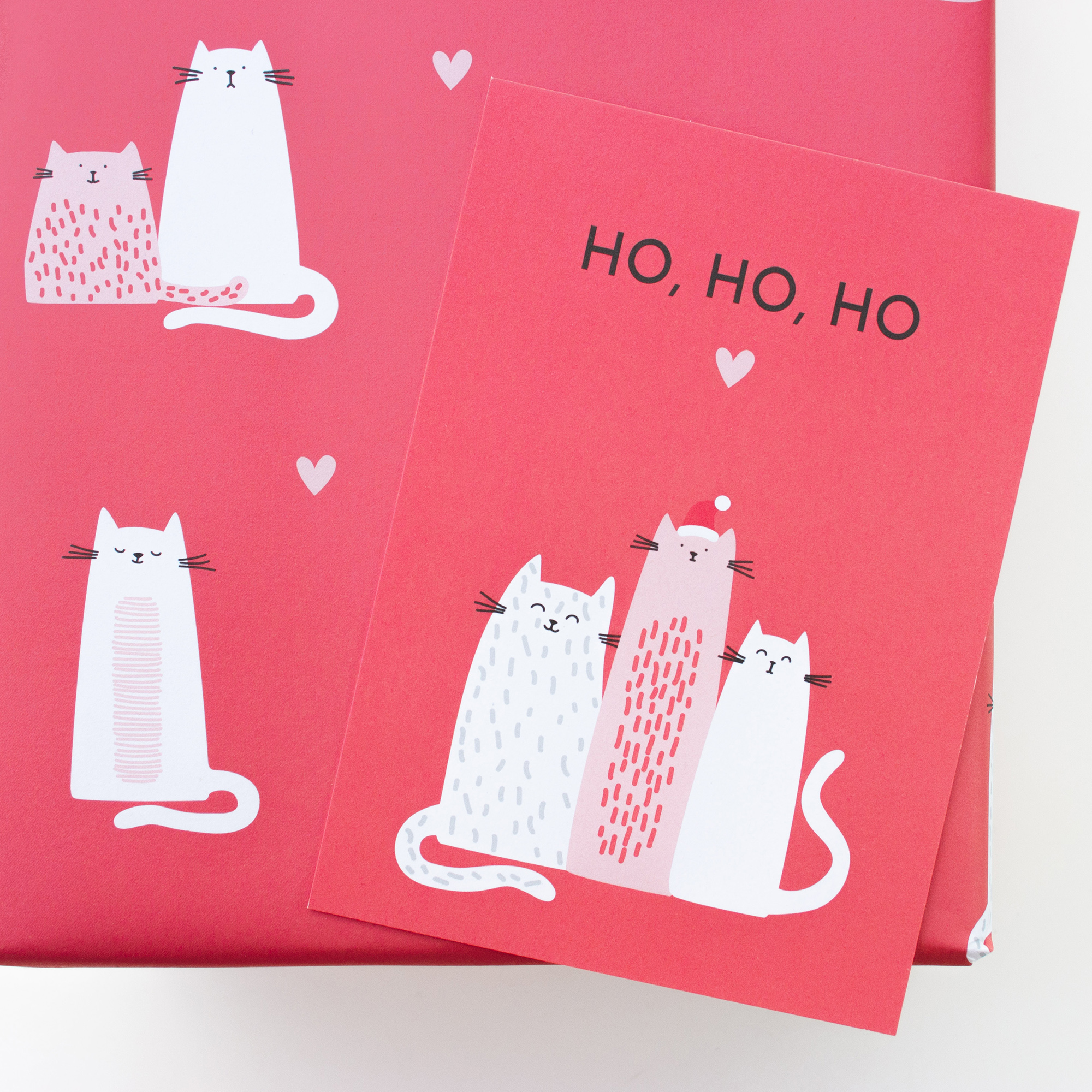 Geschenk in korallrotem Weihnachtspapier mit Katzen und Herzen, darauf die passende Katzenkarte mit dem Schriftzug „Ho, ho, ho“.