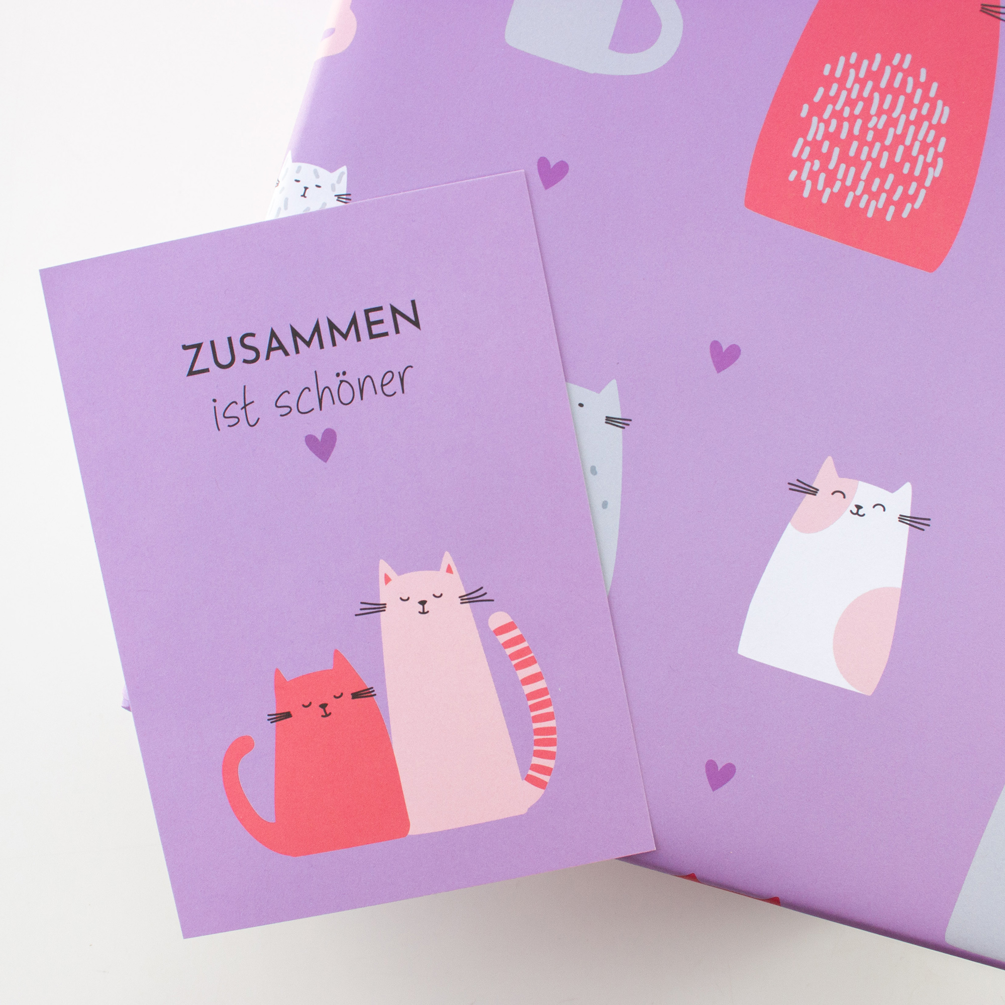 Geschenk in fliederfarbenem Geschenkpapier mit Katzen und Herzen, dazu die Katzenpostkarte „Zusammen ist schöner“.