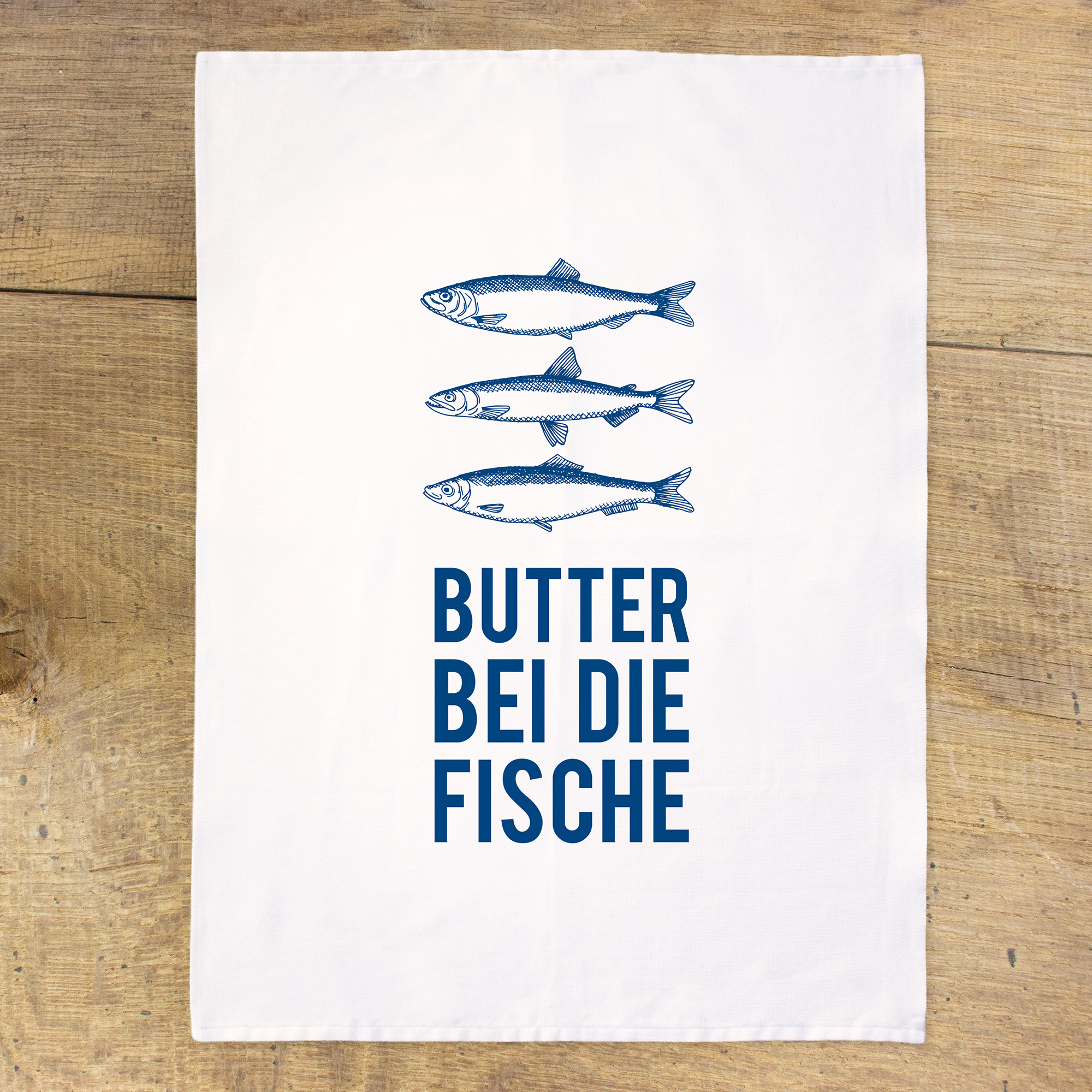 Weißes Geschirrtuch mit drei blauen Fischen über dem Spruch „Butter bei die Fische“, flach auf rustikalen Holzbrettern ausgebreitet.