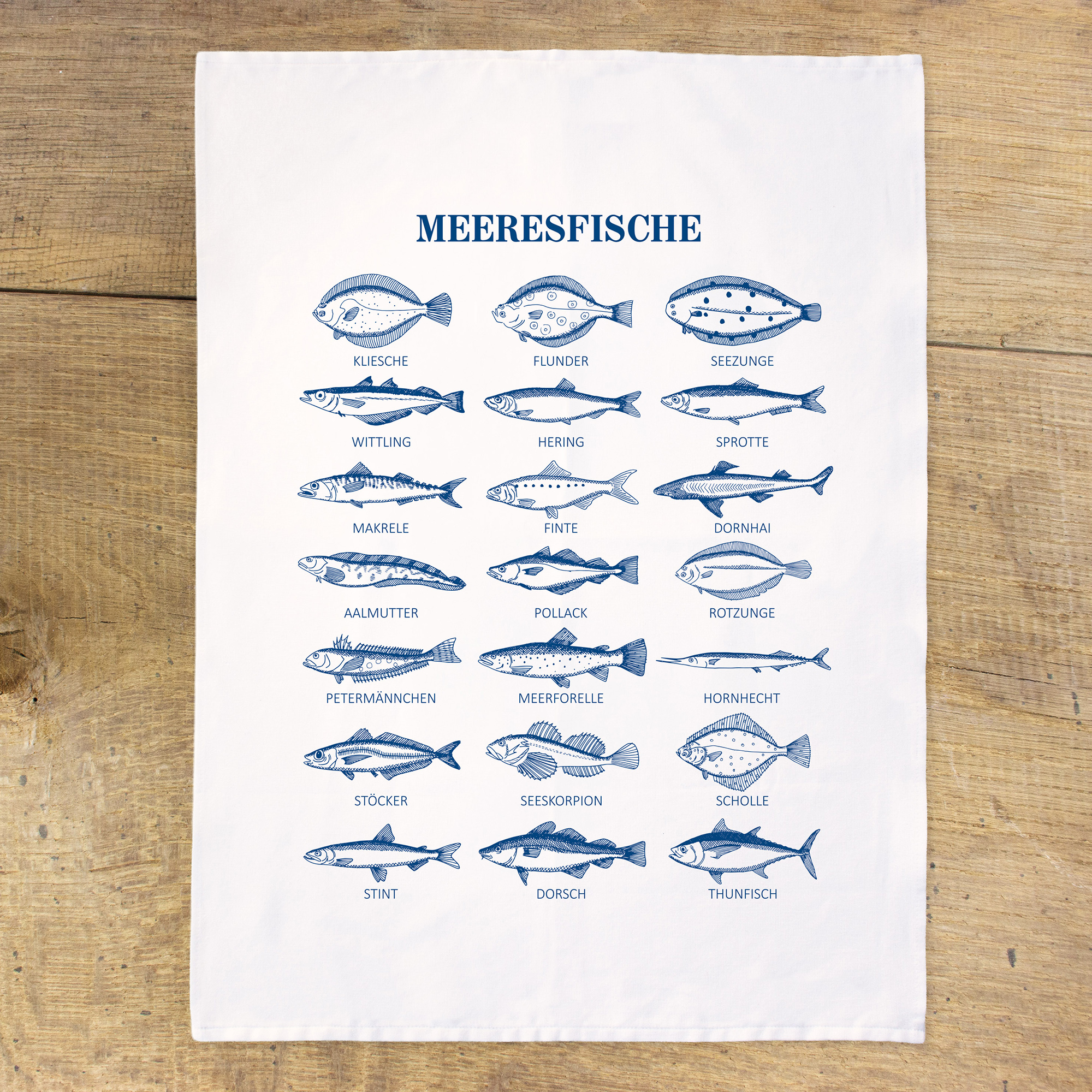 Weißes Geschirrtuch mit drei blauen Fischen über dem Spruch „Butter bei die Fische“, flach auf rustikalen Holzbrettern ausgebreitet.