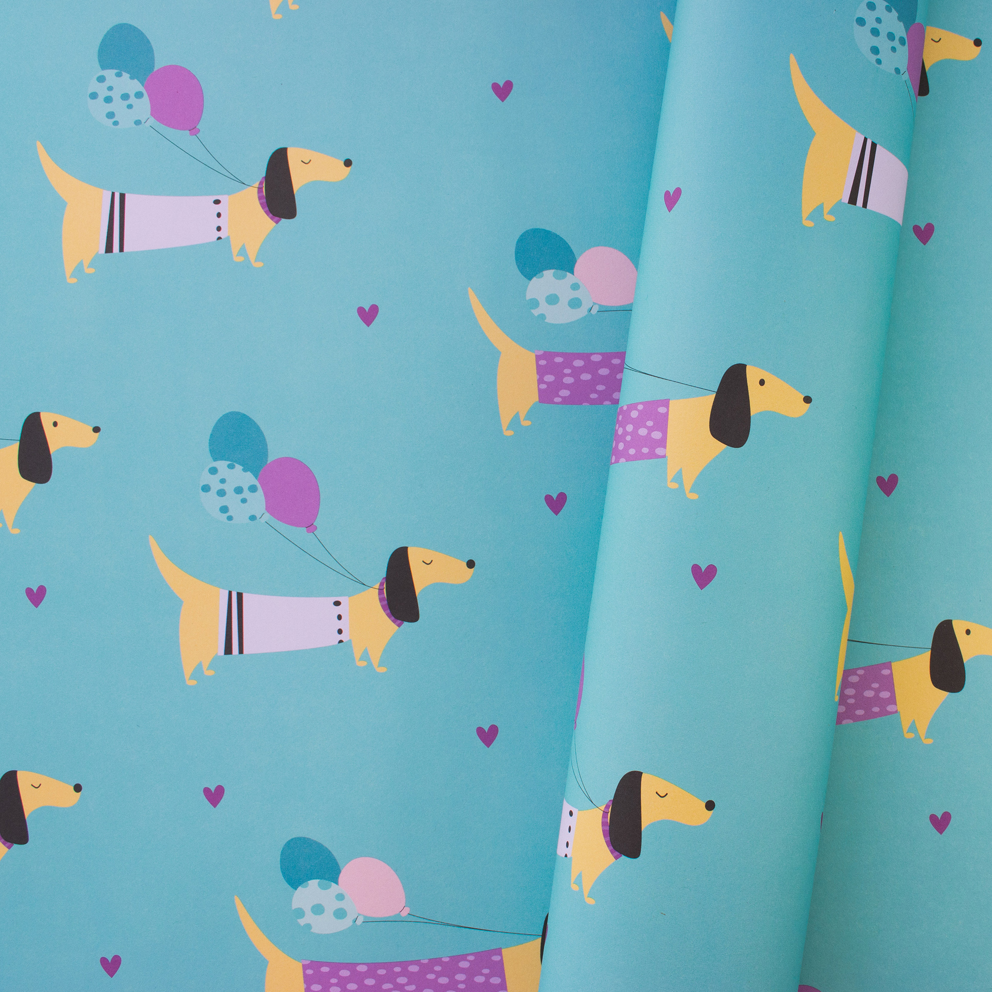 Teilweise eingerolltes Geschenkpapier „Party Dogs“ mit gelben Dackeln, bunten Luftballons und violetten Herzen auf türkisem Hintergrund.